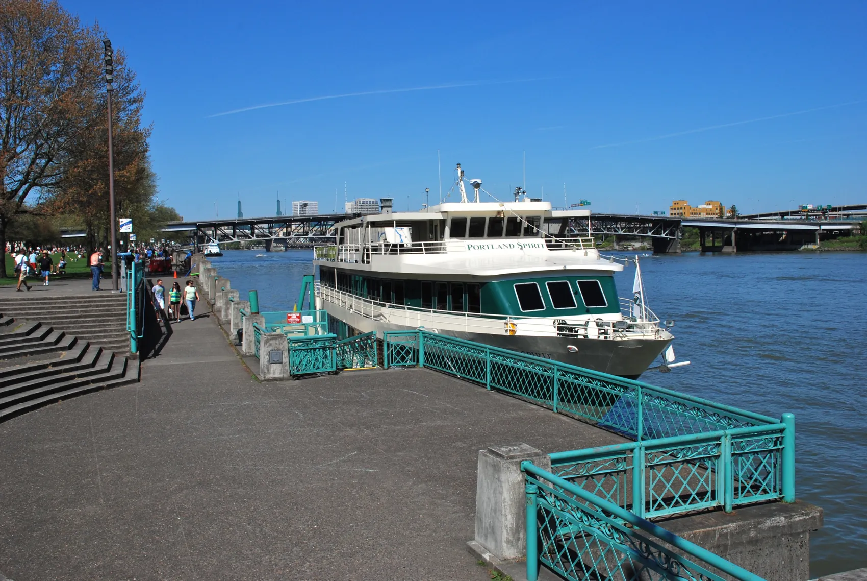 A Portland Spirit boat moored at the Waterfront Park dock beside concrete steps and a teal railing under a blue sky