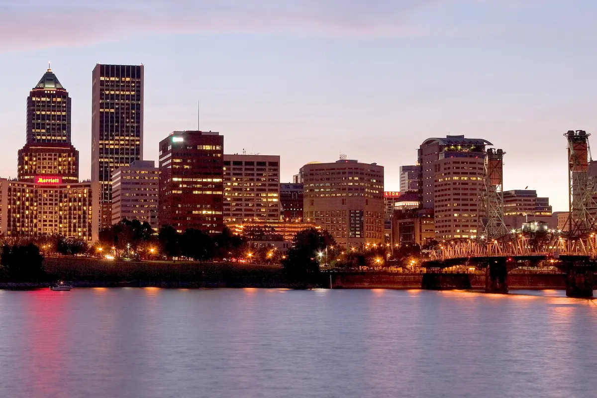 Downtown Portland towers lit at dusk above the calm Willamette River, with the Hawthorne Bridge at the right edge