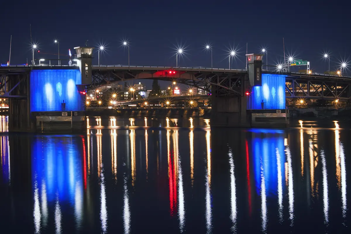 The Morrison Bridge at night, its two concrete piers lit blue and its roadway lined with bright lamps, reflected in the river